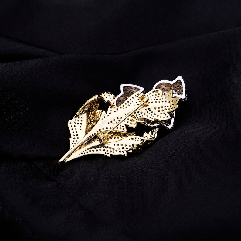 May include: A gold-tone leaf-shaped brooch with clear and black rhinestones. The brooch is set on a black fabric background.