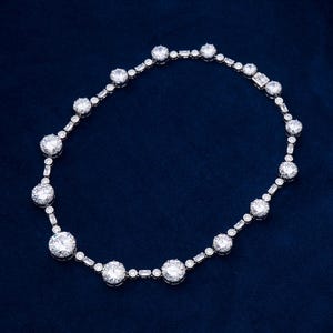May include: A silver necklace with a series of large, round, clear gemstones interspersed with smaller, rectangular and round stones. The necklace is displayed on a dark blue background, showcasing its sparkle and design.