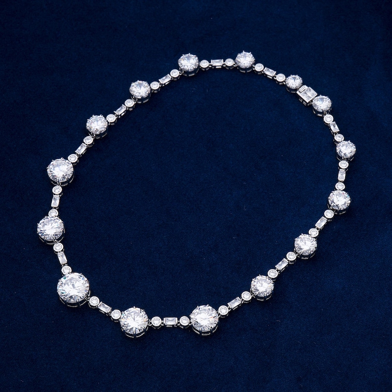 May include: A silver necklace with a series of large, round, clear gemstones interspersed with smaller, rectangular and round stones. The necklace is displayed on a dark blue background, showcasing its sparkle and design.