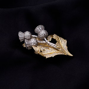 May include: A gold and silver brooch featuring a cluster of three thistle flowers with diamond accents. The thistle flowers are set against a background of gold leaves, also adorned with diamonds.