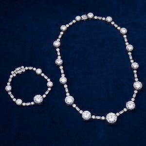 May include: A dazzling jewelry set featuring a necklace and bracelet. The necklace and bracelet are adorned with large, round, and rectangular-cut clear gemstones, set in a silver-toned metal. The pieces are displayed on a dark blue background.