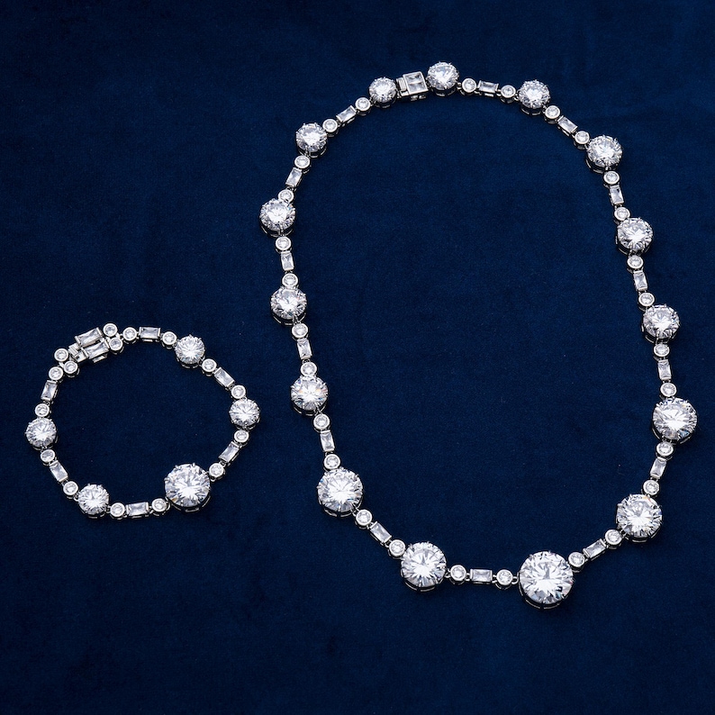 May include: A dazzling jewelry set featuring a necklace and bracelet. The necklace and bracelet are adorned with large, round, and rectangular-cut clear gemstones, set in a silver-toned metal. The pieces are displayed on a dark blue background.
