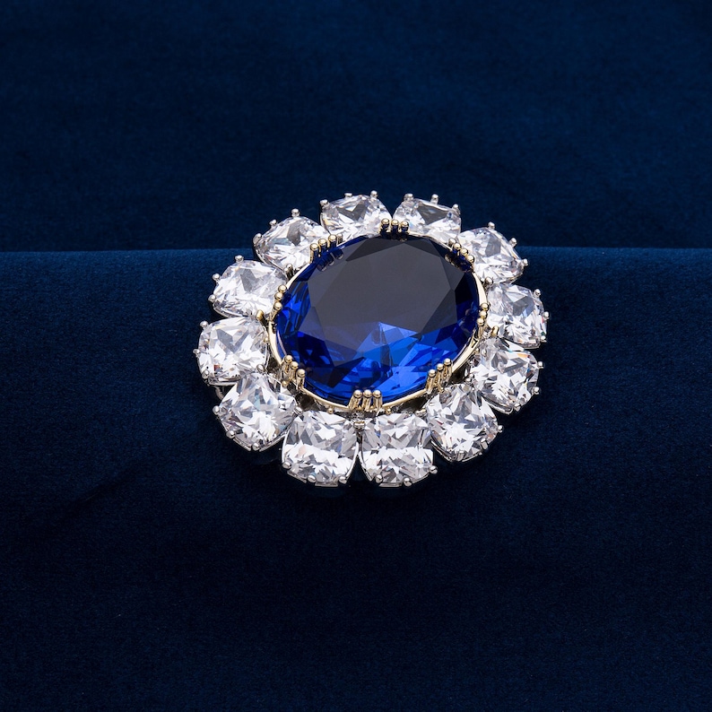 May include: A dazzling brooch featuring a large, oval, deep blue gemstone at its center, surrounded by a halo of sparkling, clear, square-cut stones. The setting is gold-toned, and the brooch is set against a dark blue background.