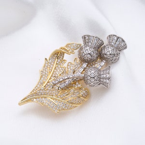 May include: A gold and silver brooch featuring a cluster of three thistle flowers with sparkling clear rhinestones. The thistle flowers are set against a gold leaf design.