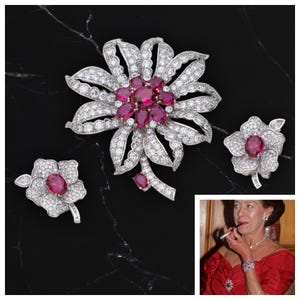 May include: A silver-toned floral jewellery set comprising a large brooch and two smaller pieces. The brooch and earrings are adorned with clear crystals and feature a cluster of oval-cut, deep red gemstones. The set is displayed on a black surface.