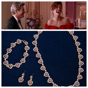 以下が含まれることがあります： 赤いハート型の石がついた金のネックレス、ブレスレット、イヤリングのセット。ジュエリーは青い背景の上にあります。