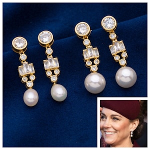 May include: Gold-toned drop earrings with a large white pearl at the bottom. The earrings feature a round, clear stone at the top, rectangular and round clear stones in the middle, and are set against a dark blue background. The earrings are shown in a close-up shot.