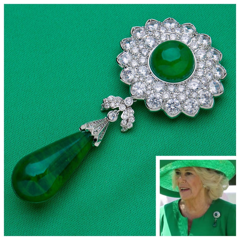 May include: A large, ornate brooch with a central green gemstone surrounded by clear, sparkling stones. A teardrop-shaped green gemstone hangs below, connected by a detailed silver-toned design. The brooch is set against a solid green background.