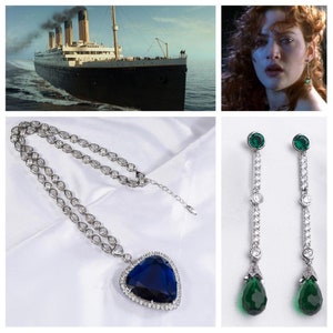 Peut inclure: Un collier en argent avec un grand pendentif en forme de cœur bleu entouré de petits diamants. Le pendentif est suspendu à une chaîne en argent avec de petits diamants. Le collier est sur un fond blanc. Une paire de boucles d'oreilles en argent avec une pierre verte en forme de larme suspendue à une chaîne en argent avec de petits diamants.