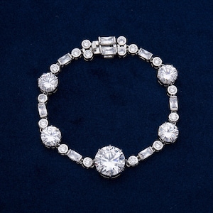 May include: A silver bracelet with a series of round and rectangular clear stones. The bracelet features a clasp closure and is set against a dark blue background. The stones are arranged in a repeating pattern.