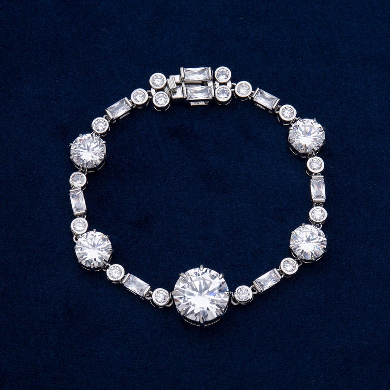 May include: A silver bracelet with a series of round and rectangular clear stones. The bracelet features a clasp closure and is set against a dark blue background. The stones are arranged in a repeating pattern.