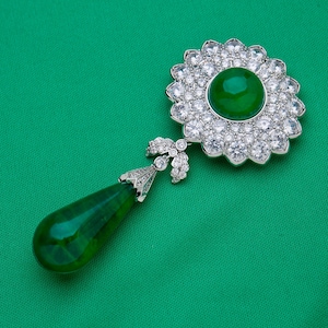 May include: A detailed view of a brooch with a large, round, emerald-green gemstone at the center, surrounded by a halo of sparkling clear stones. A teardrop-shaped green stone hangs below, connected by a decorative silver-toned element.