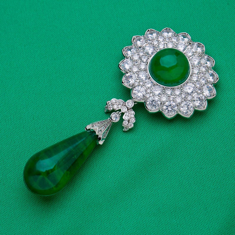 May include: A detailed view of a brooch with a large, round, emerald-green gemstone at the center, surrounded by a halo of sparkling clear stones. A teardrop-shaped green stone hangs below, connected by a decorative silver-toned element.