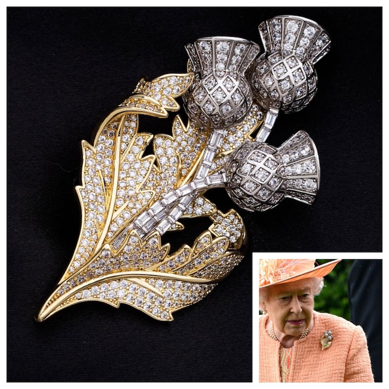 May include: A gold and silver brooch featuring a cluster of three thistle flowers, each adorned with sparkling clear rhinestones. The thistle flowers are set on a leafy stem, also encrusted with rhinestones.
