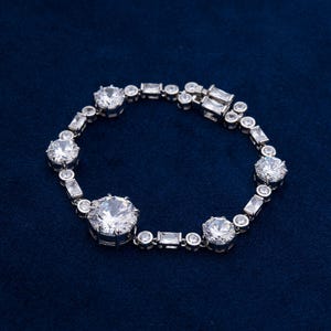 May include: A silver bracelet with multiple clear, round, and rectangular-cut stones. The bracelet features a series of large, round stones interspersed with smaller, rectangular and round stones, all set in a silver metal band. The bracelet is displayed on a dark blue surface.