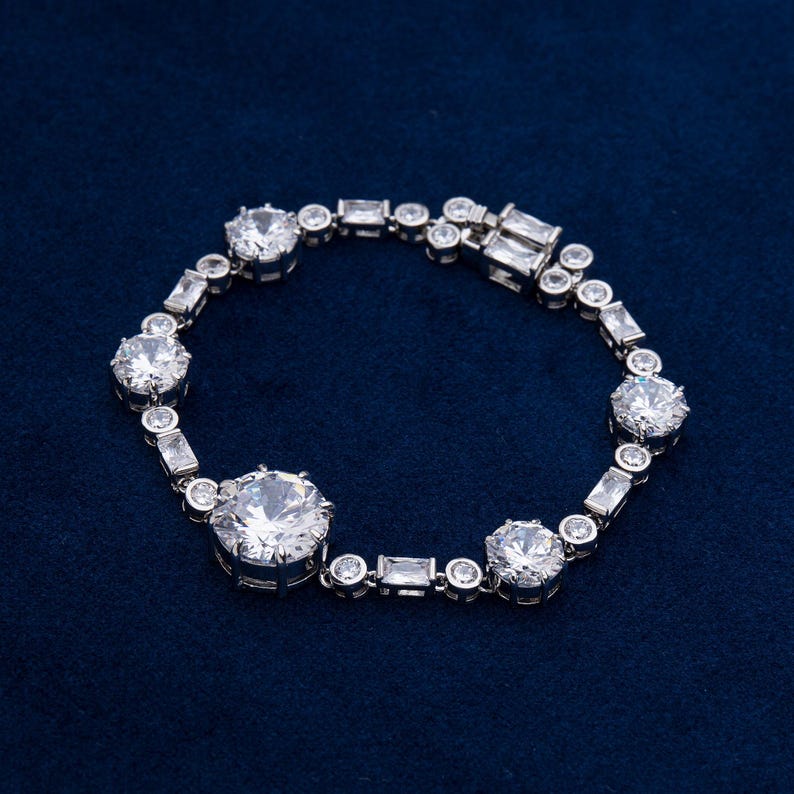 May include: A silver bracelet with multiple clear, round, and rectangular-cut stones. The bracelet features a series of large, round stones interspersed with smaller, rectangular and round stones, all set in a silver metal band. The bracelet is displayed on a dark blue surface.