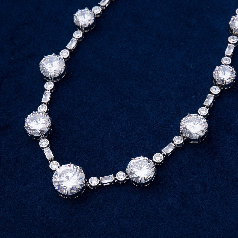 May include: A silver necklace with large, round, clear gemstones and smaller, rectangular and round gemstones. The necklace is displayed on a dark blue background. The gemstones are set in a silver metal.