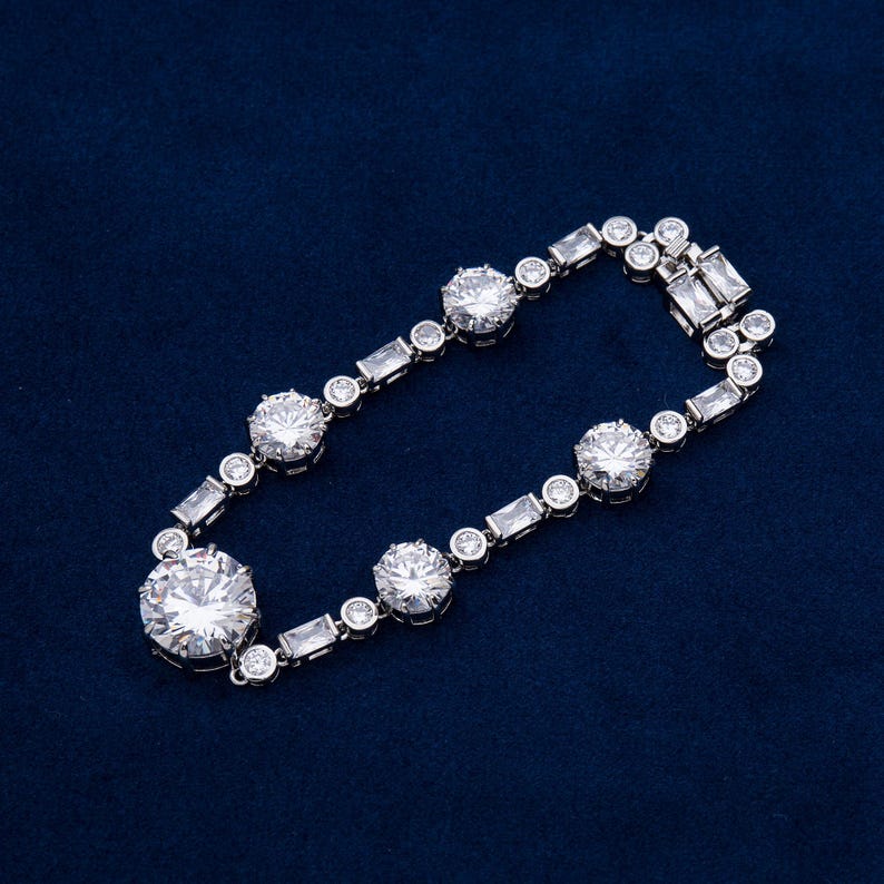 May include: A silver bracelet with large, round, clear gemstones and smaller, rectangular and round stones. The bracelet is set against a dark blue background, showcasing its sparkle and design.