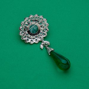 May include: A silver-toned brooch with a floral design, featuring a central teal stone and surrounding clear crystals. A green teardrop-shaped pendant hangs below. The brooch is set against a green background.