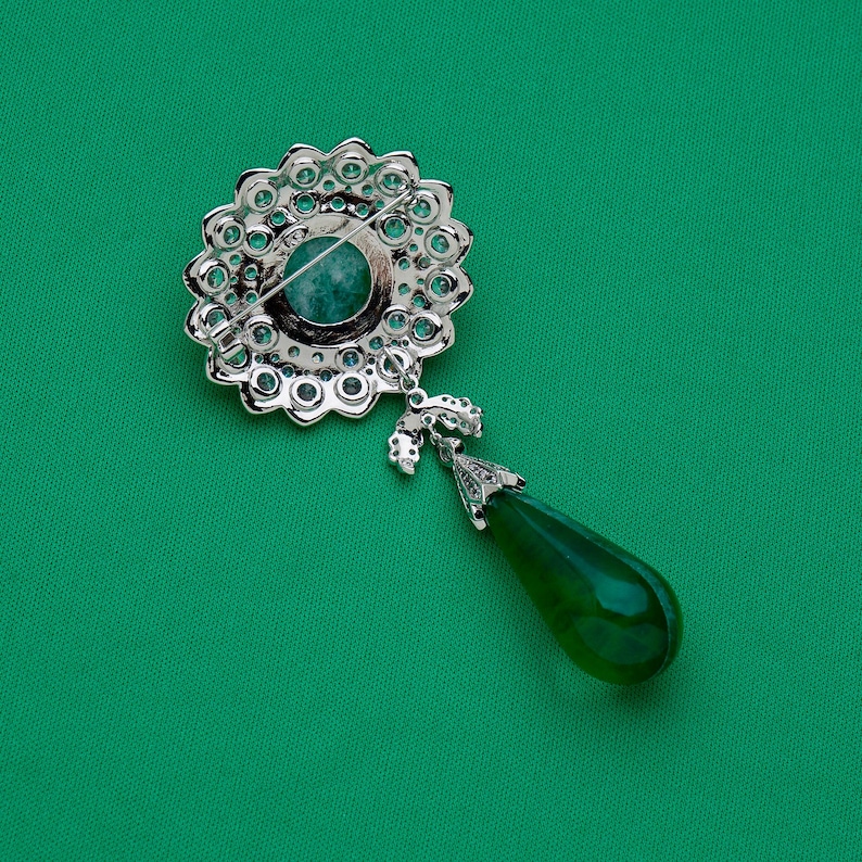 May include: A silver-toned brooch with a floral design, featuring a central teal stone and surrounding clear crystals. A green teardrop-shaped pendant hangs below. The brooch is set against a green background.