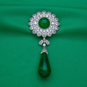 May include: A silver brooch with a large, round, emerald-green center stone surrounded by a halo of clear, sparkling crystals. A teardrop-shaped green stone hangs below. The brooch is set against a green background.