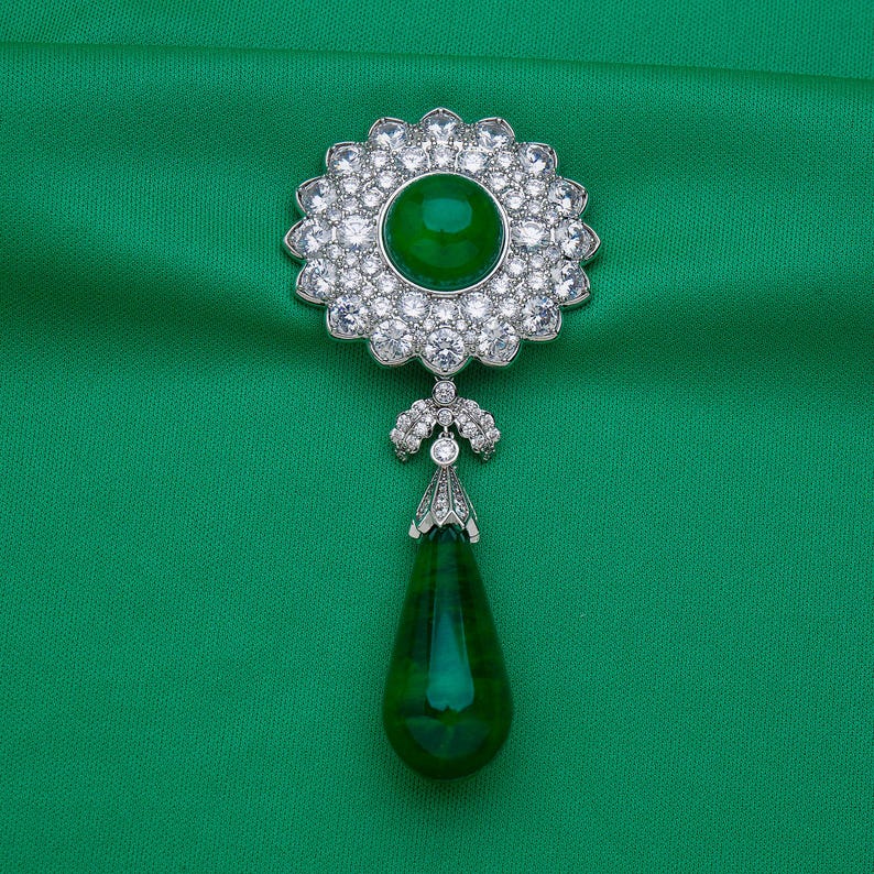 May include: A silver brooch with a large, round, emerald-green center stone surrounded by a halo of clear, sparkling crystals. A teardrop-shaped green stone hangs below. The brooch is set against a green background.