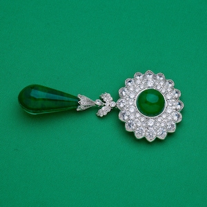 May include: A pendant featuring a large, round, emerald-green gemstone surrounded by a halo of clear, sparkling stones. A teardrop-shaped green stone hangs from the pendant, connected by a detailed, silver-colored metal design.