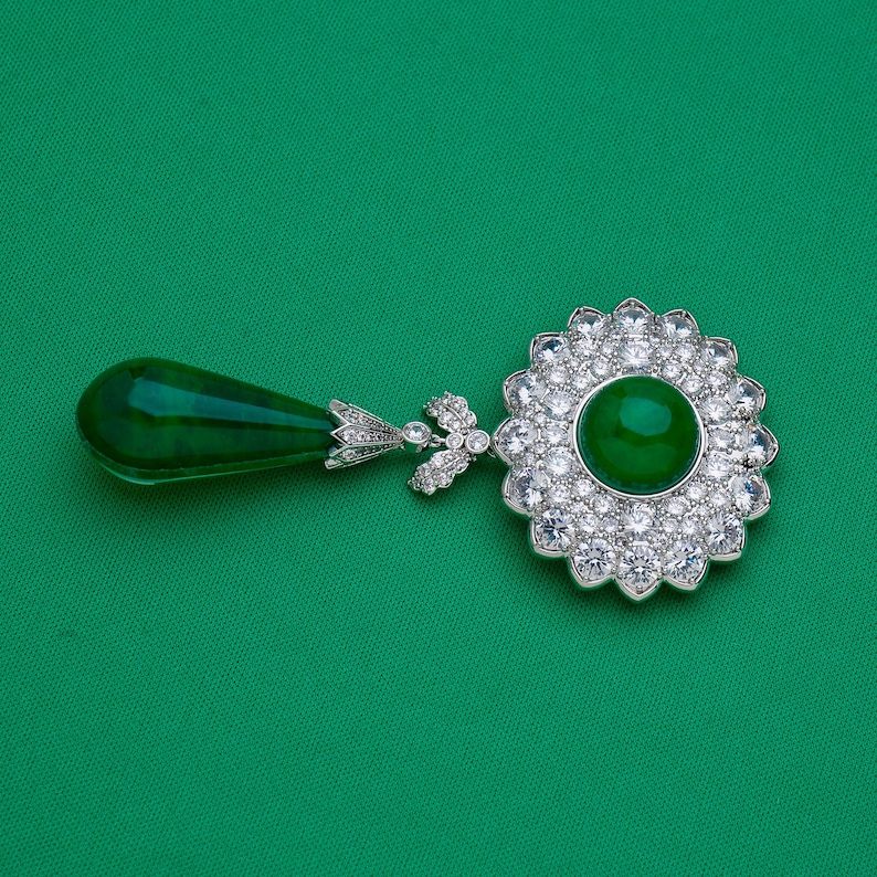 May include: A pendant featuring a large, round, emerald-green gemstone surrounded by a halo of clear, sparkling stones. A teardrop-shaped green stone hangs from the pendant, connected by a detailed, silver-colored metal design.