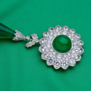 May include: A silver pendant with a large, round, vibrant green gemstone center, surrounded by a halo of smaller, clear gemstones. The pendant has a floral design and a matching green teardrop-shaped accent.