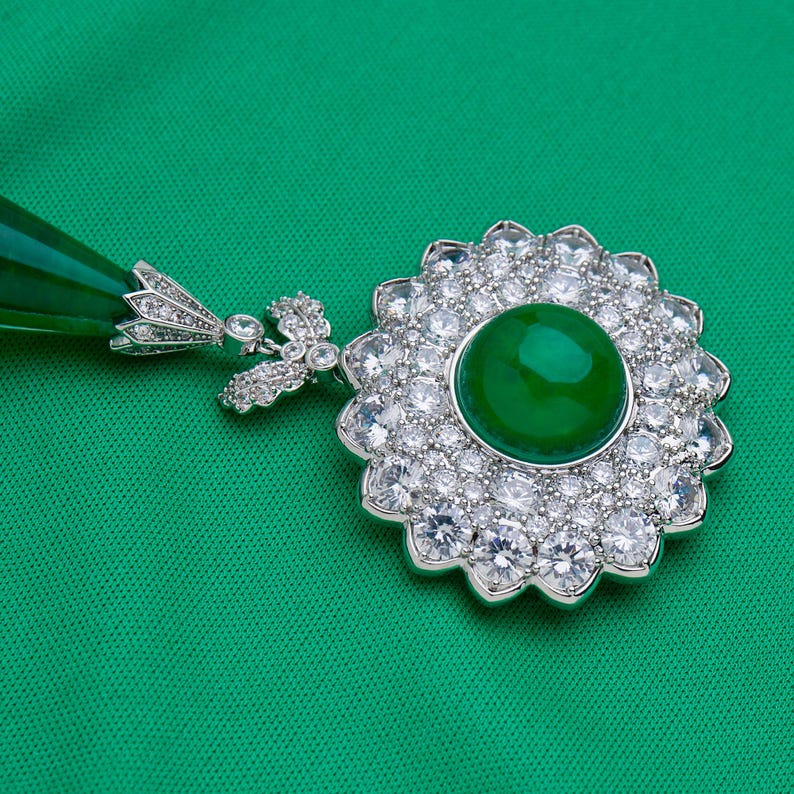 May include: A silver pendant with a large, round, vibrant green gemstone center, surrounded by a halo of smaller, clear gemstones. The pendant has a floral design and a matching green teardrop-shaped accent.