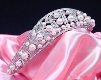 May include: A silver tiara with a delicate design featuring faux pearls and rhinestones. The tiara is curved and rests on a pink satin fabric.