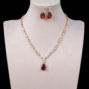 May include: A gold-coloured jewellery set comprising a necklace and matching earrings. The necklace and earrings feature teardrop-shaped, deep red gemstones. The necklace has a delicate chain with clear, sparkling accents. The earrings dangle from small, clear stones.