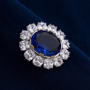 May include: A large, oval-shaped blue gemstone is the focal point of this brooch, surrounded by a halo of square, clear gemstones. The brooch has a gold-colored setting and is set against a dark blue velvet background.