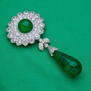 May include: A detailed shot of a brooch with a large, round, vibrant green center stone surrounded by a halo of smaller, sparkling clear stones. A teardrop-shaped green stone hangs below. The brooch is set against a green background.