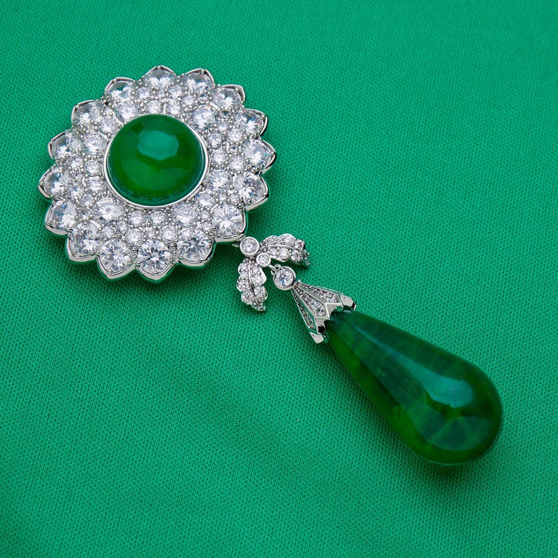 May include: A detailed shot of a brooch with a large, round, vibrant green center stone surrounded by a halo of smaller, sparkling clear stones. A teardrop-shaped green stone hangs below. The brooch is set against a green background.