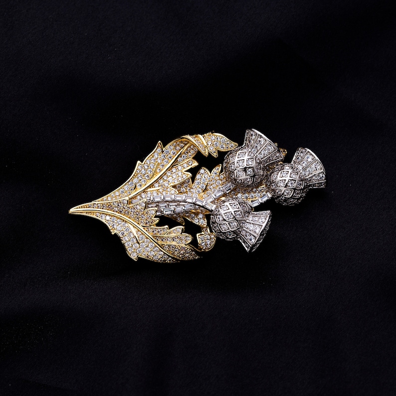 May include: A gold and silver brooch with a thistle design. The brooch is adorned with sparkling clear rhinestones.