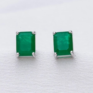 May include: A pair of emerald-cut, bright green gemstone stud earrings. The gemstones are set in a silver-coloured metal with four prongs. The earrings are displayed on a white, textured surface.