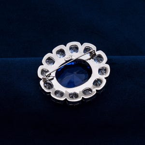 May include: A silver-toned brooch with a large, oval, deep blue gemstone at its center, surrounded by smaller, clear gemstones. The brooch has a pin closure on the back and a scalloped edge. The background is a dark blue.