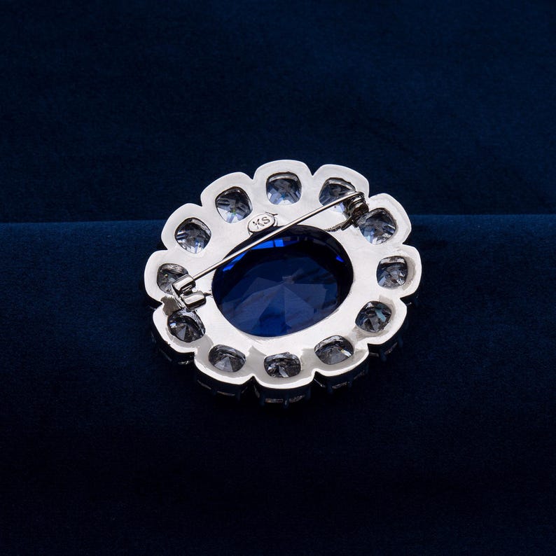 May include: A silver-toned brooch with a large, oval, deep blue gemstone at its center, surrounded by smaller, clear gemstones. The brooch has a pin closure on the back and a scalloped edge. The background is a dark blue.