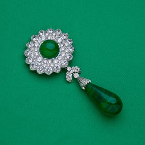 May include: A silver-toned brooch with a large, round, emerald-green center stone surrounded by clear, sparkling crystals. A teardrop-shaped green stone hangs below, connected by a decorative silver element. The brooch is set against a green background.