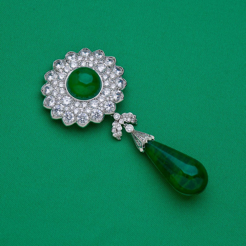 May include: A silver-toned brooch with a large, round, emerald-green center stone surrounded by clear, sparkling crystals. A teardrop-shaped green stone hangs below, connected by a decorative silver element. The brooch is set against a green background.