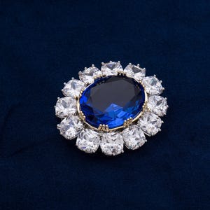 May include: A dazzling brooch featuring a large, oval, deep blue gemstone at the center, surrounded by a halo of square, clear crystals. The setting has a gold-colored rim, creating a striking contrast against the dark blue background.