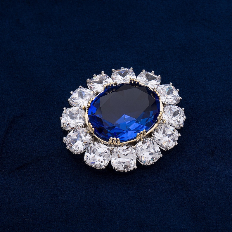 May include: A dazzling brooch featuring a large, oval, deep blue gemstone at the center, surrounded by a halo of square, clear crystals. The setting has a gold-colored rim, creating a striking contrast against the dark blue background.