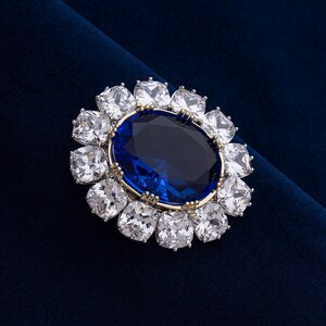 May include: A large, oval-shaped blue gemstone is the focal point of this brooch, surrounded by a halo of square, clear gemstones. The brooch has a gold-colored setting and is set against a dark blue velvet background.