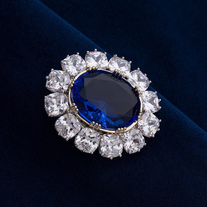 May include: A large, oval-shaped blue gemstone is the focal point of this brooch, surrounded by a halo of square, clear gemstones. The brooch has a gold-colored setting and is set against a dark blue velvet background.