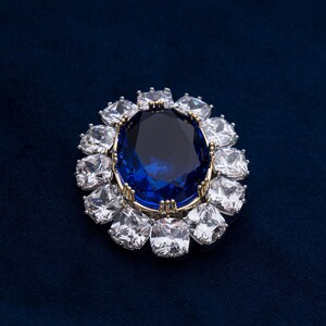 May include: A gold-toned brooch featuring a large, oval, deep blue gemstone surrounded by smaller, square, clear gemstones. The brooch is set against a dark blue background, creating a striking contrast.