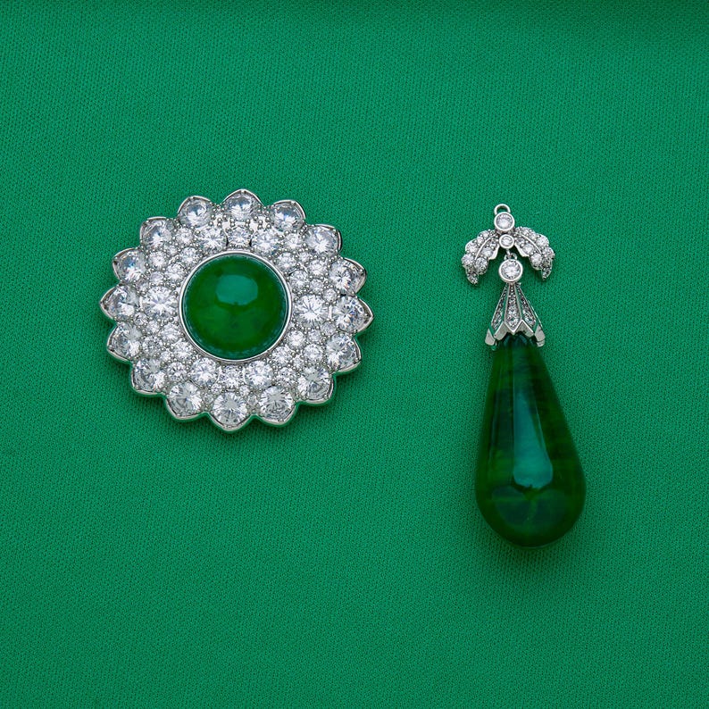 May include: A floral-shaped brooch and a teardrop pendant, both featuring a vibrant green center stone surrounded by sparkling clear crystals. The jewelry is set against a solid green background.