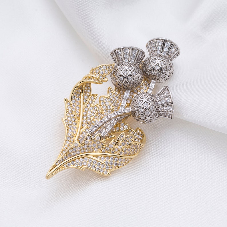 May include: A gold and silver brooch featuring a stylized thistle design with clear rhinestones. The brooch is a traditional Scottish symbol.