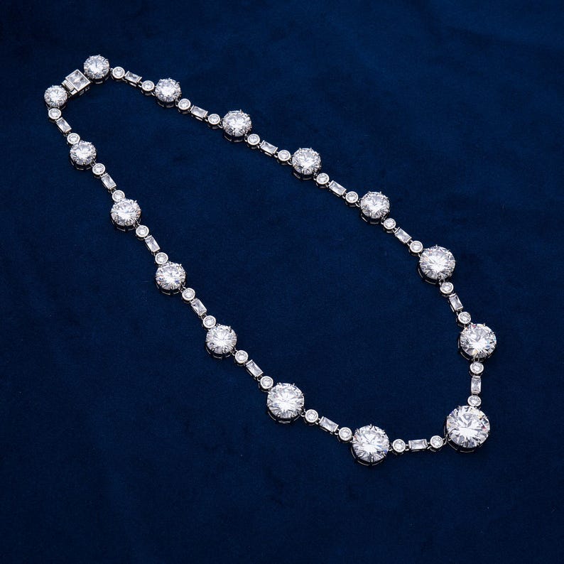 May include: A dazzling necklace featuring a series of large, round, clear gemstones interspersed with smaller, rectangular and round stones. The necklace is set in a silver-toned metal and rests against a deep blue background, creating a striking contrast.