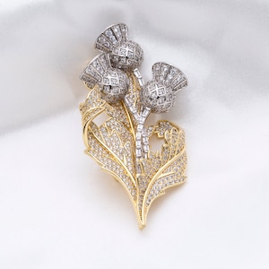 May include: A gold and silver brooch featuring a cluster of three thistle flowers with sparkling rhinestones. The thistle flowers are set against a backdrop of gold leaves, also adorned with rhinestones.